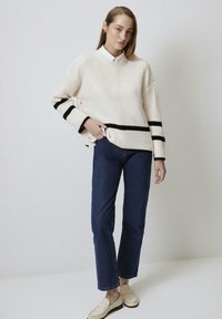 Pull en maille beige à rayures noires, avec des boutons dorés sur le côté. Assorti à un jean droit bleu foncé et des mocassins clairs.