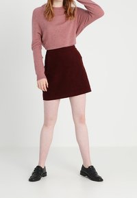 Bordowa mini spódnica z korduroy w zestawieniu z jasnoróżowym swetrem o teksturowej fakturze. Czarne buty wsuwane dopełniają całość.