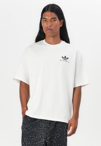 Baltas kokvilnas T-krekls ar īsām piedurknēm, ar melnu izdrukātu Adidas basketbola logotipu uz krūtīm. Gluda tekstūra, atbrīvota piegriezums.