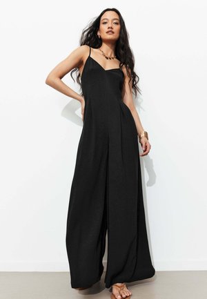 Femme aux longs cheveux foncés portant une combinaison noire sans manches à jambes larges et des bijoux dorés, debout contre un mur blanc.