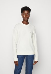 White knit sweater with long sleeves and ribbed cuffs. Features a small green logo on the left chest. Paired with blue jeans.