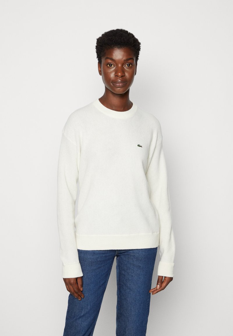 White knit sweater with long sleeves and ribbed cuffs. Features a small green logo on the left chest. Paired with blue jeans.
