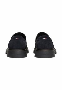 Granatowe zamszowe buty wsuwane na czarnym gumowym spodzie, z małym emblematem w kolorach czerwonym, białym i niebieskim z tyłu. Minimalistyczny design.