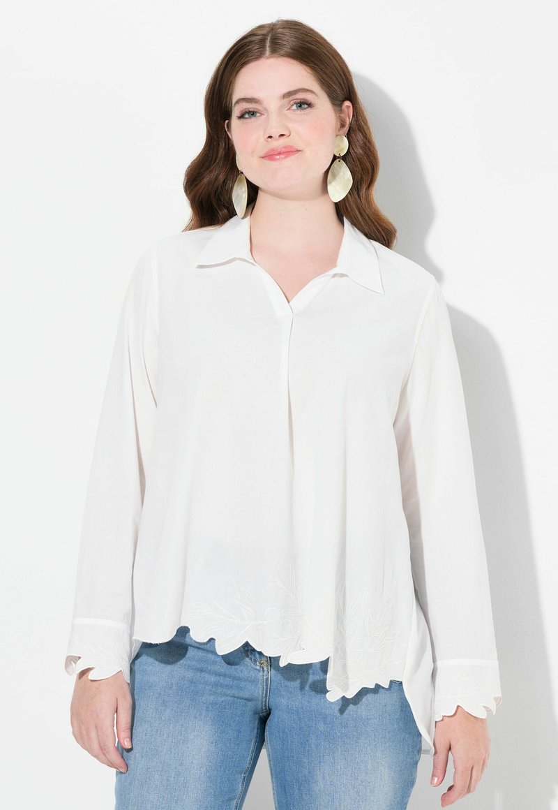 Blusa bianca a maniche lunghe con colletto e orlo scallopato, caratterizzata da dettagli ricamati a foglia. Abbinata a jeans blu.
