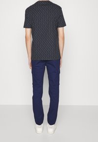 Osoba stojící zády k fotoaparátu, oblečená v černém tričku s krátkým rukávem a bílými tečkami, tmavě modrých cargo kalhotách a bílých teniskách Veja na bílém pozadí.