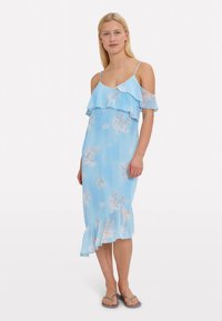 Egy hölgy világoskék virágmintás, hideg vállú midi ruhában áll, amely fodrokkal díszített, és szürke szandálokat visel, háttérben egy fehér háttérrel.