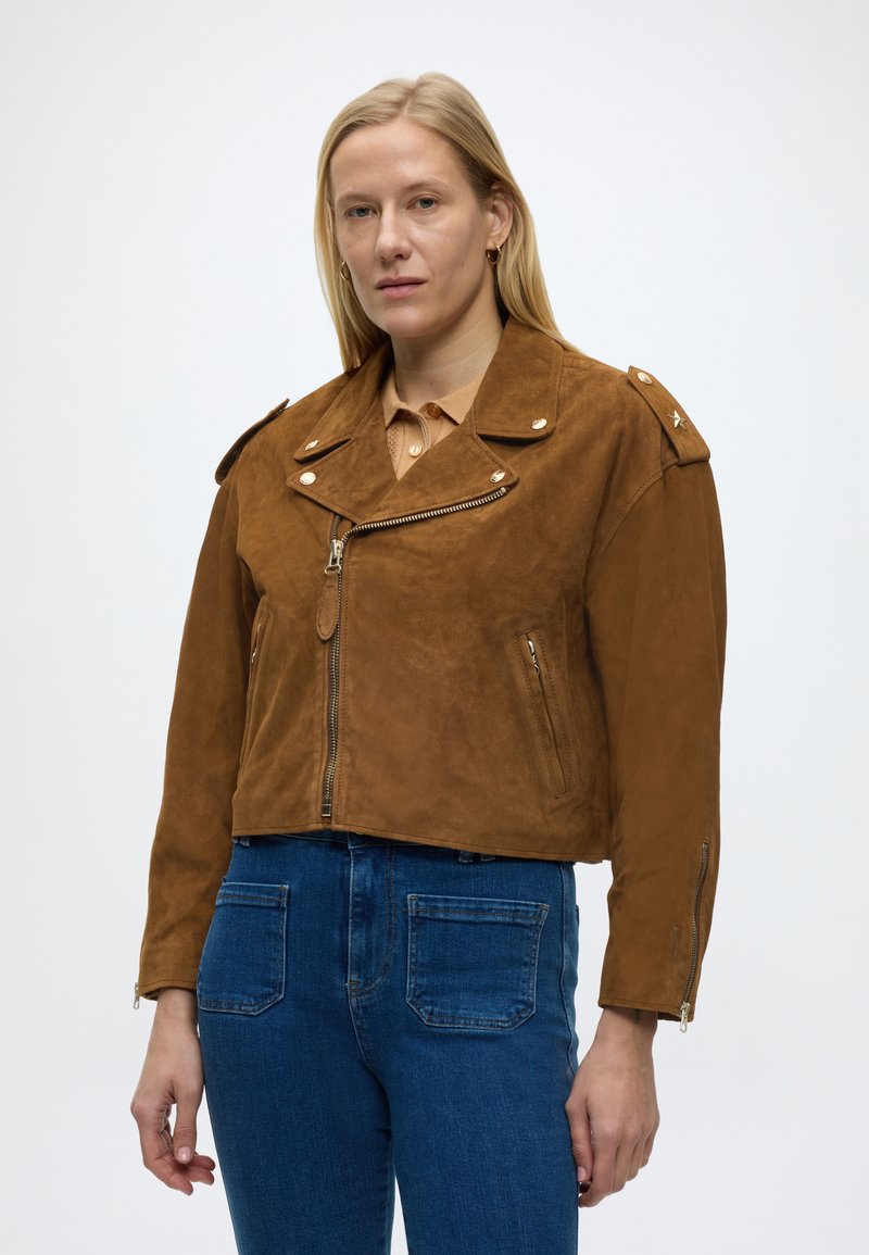 Γυναίκα με ίσια ξανθά μαλλιά που φοράει καφέ σατέν δερμάτινο μπουφάν τύπου μοτοσικλετιστή και μπλε τζιν, στέκεται μπροστά σε απλό λευκό φόντο.