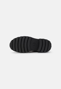 Suela de zapato de goma negra con ranuras horizontales, que presenta una puntera redonda y una superficie texturizada. Incluye un detalle del logo circular.
