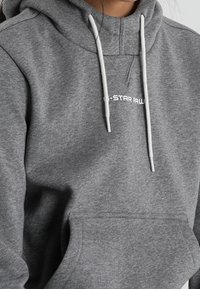 Sudadera gris con bolsillo frontal y cordones, con un logo en texto blanco "C-STAR RAW" en la zona del pecho. Textura de tela suave.
