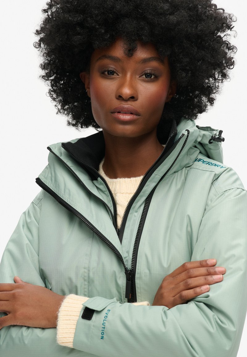 Lichtgroene waterdichte jas met een hoge kraag, ritssluiting en zwarte accenten. Beschikt over manchetten met verstelbare klittenbandsluiting en merkdetails.