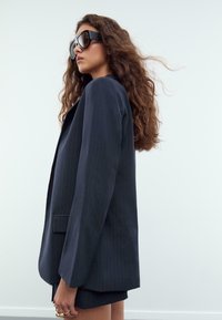 Kvinna med långt lockigt hår iförd en oversized svart rutiga blazer, stora svarta solglasögon och guldringar, stående mot en enkel vit bakgrund.