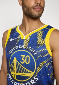 Golden State Warriors-drakt i blå og gul. Har mesh-stoff, V-hals-design, sirkulært logo med bro-grafikk og spiller nummer.