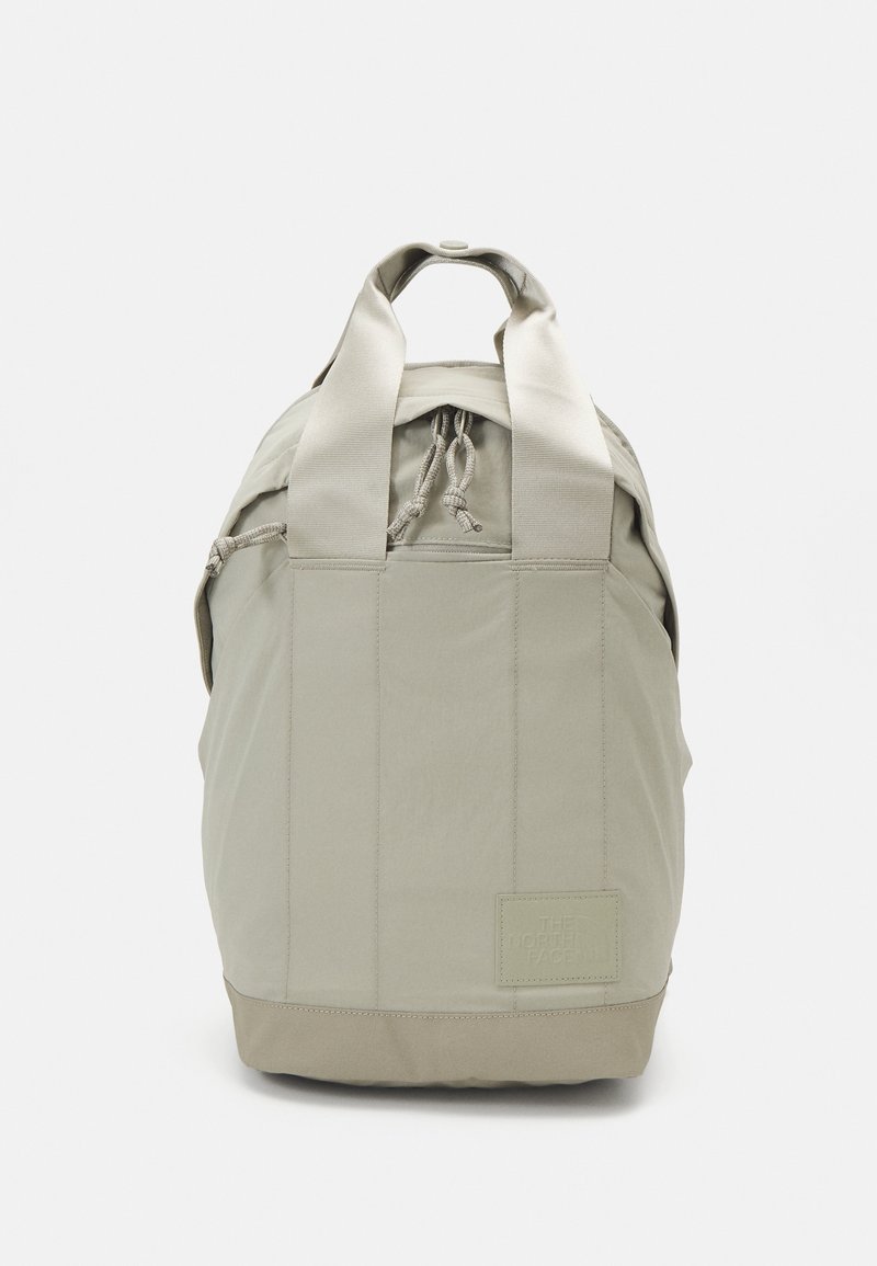 Sac à dos beige avec double poignée de transport, fermeture par cordon et un logo patch carré sur le bas à droite à l'avant.