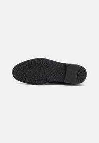 Semelle de chaussure noire avec une bande de roulement texturée et à motif, conçue pour l'adhérence et la durabilité, montrée sur un fond clair uni.