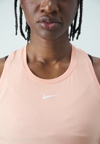 Top sportiv Nike în piersică deschisă, cu guler rotund și design racerback. Materialul pare neted, având un mic logo alb pe piept.