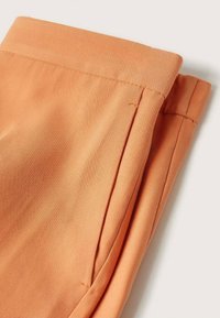 Gros plan d'un pantalon orange plié montrant la taille, la couture latérale et la surpiqûre des poches sur un fond uni clair.