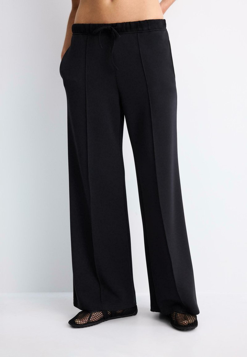 Personne portant un pantalon noir à jambes larges avec coutures devant, taille à cordon, mains dans les poches et chaussures noires en mesh sans lacets.
