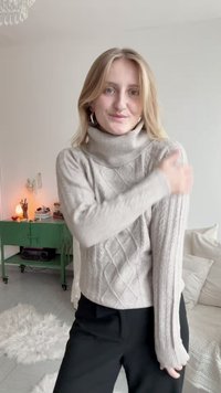 Vaaleanharmaa kaapelkudottu kauluspaita, jossa on ribbauskoristeita, pitkät hihat ja tiukka siluetti, yhdistetty mustiin housuihin.