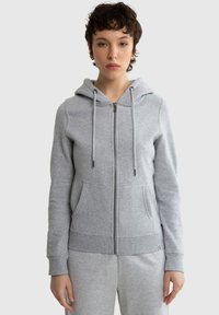 Noor inimene kannab helehallist lukuga kapuutsiga pusa, millel on ees taskud, ja sobivaid halli värvi dressipükse.