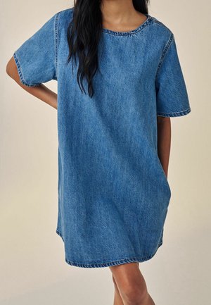 Personne portant une robe en jean ample à manches courtes, debout avec une main sur la hanche devant un fond uni clair. Le visage n'est pas visible.