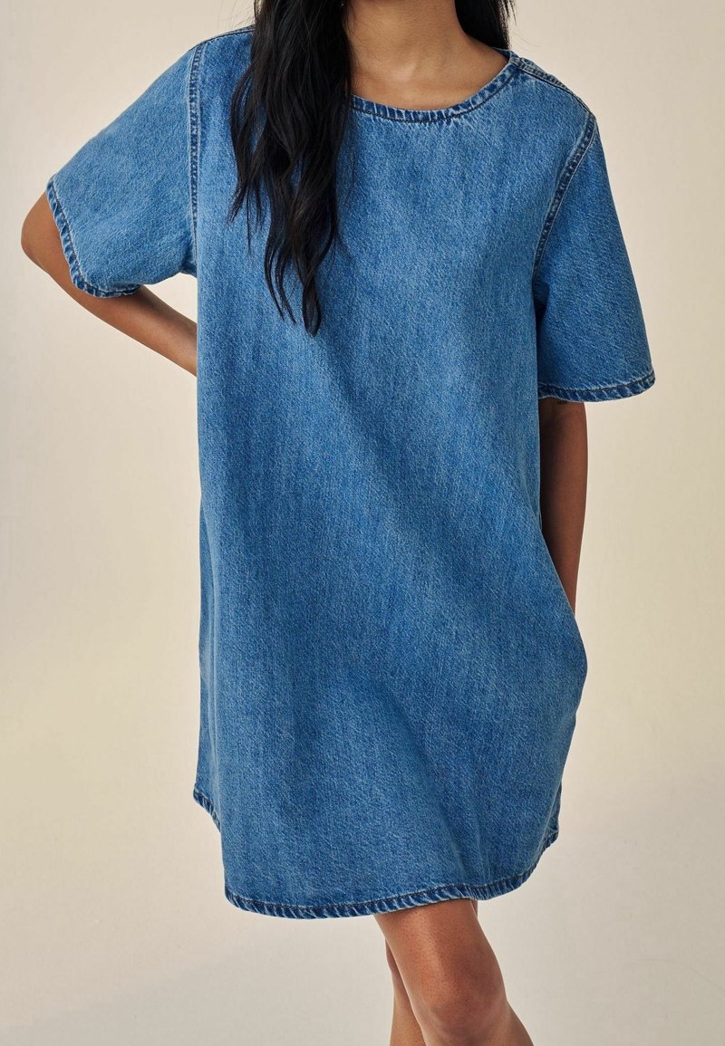 Personne portant une robe en jean ample à manches courtes, debout avec une main sur la hanche devant un fond uni clair. Le visage n'est pas visible.