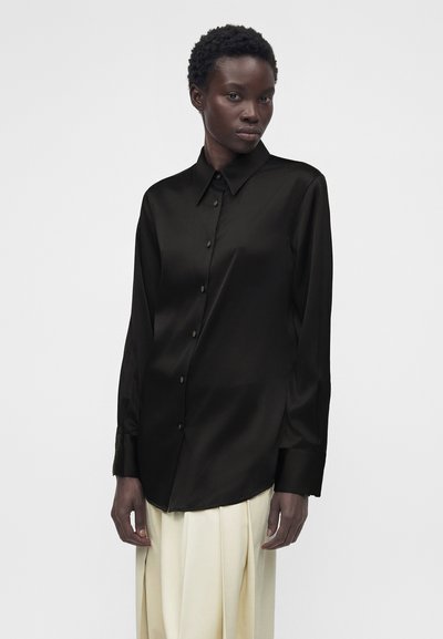 Γυναίκα που φοράει μαύρο σατέν πουκάμισο με κουμπιά και μακριά μανίκια και κρεμ πλισέ φούστα, με φόντο απλό ανοιχτό χρώμα.