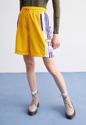 Personne portant un short en mesh jaune Adidas avec des rayures violettes, des bottes beiges à lacets et un haut vert tie-dye à manches longues, debout sur un sol bleu.