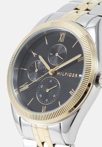 Montre en argent et or avec un cadran noir, des chiffres romains et trois sous-cadrans. Présente une lunette texturée et un bracelet en métal relié.