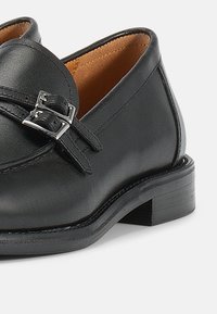 Μαύρα δερμάτινα μοκασίνια με ασημένια αγκράφα, χαμηλό σωρευμένο τακούνι και ομαλή υφή. Το εσωτερικό είναι επενδυμένο με ταμπά υλικό.