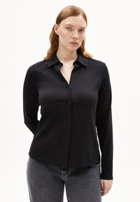 Camicia nera con bottoni, maniche lunghe, colletto e una texture liscia. Abbinata a pantaloni grigi, esposta su uno sfondo bianco.