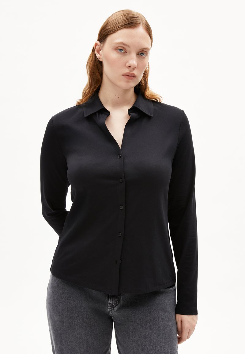 Camicia nera con bottoni, maniche lunghe, colletto e una texture liscia. Abbinata a pantaloni grigi, esposta su uno sfondo bianco.