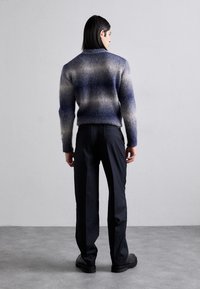 Pull en peluche pour homme en dégradé de bleu et gris avec un col haut, associé à un pantalon noir et des chaussures noires massives, vu de dos.