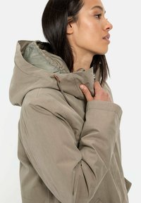 Beige Kapuzenjacke mit einer strukturierten Außenseite und einem glatten, hellen Innenfutter. Verfügt über eine verstellbare Kordelzug und seitliche Taschen.