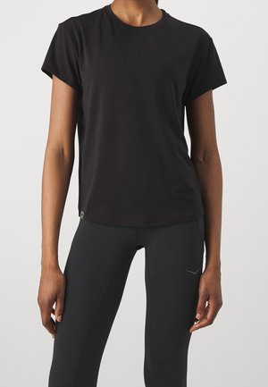Person trägt ein schlichtes schwarzes kurzärmeliges T-Shirt und schwarze Leggings, steht mit locker an den Seiten hängenden Armen vor einem schlichten Hintergrund.