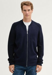 Granatowy kardigan zapinany na zamek z prążkowanym kołnierzem i mankietami, o gładkiej fakturze i dopasowanym kroju, w zestawie z białą koszulką i dżinsami.