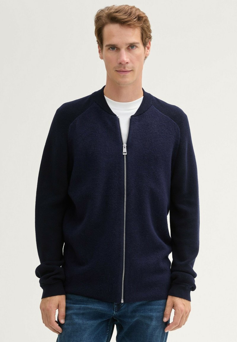 Granatowy kardigan zapinany na zamek z prążkowanym kołnierzem i mankietami, o gładkiej fakturze i dopasowanym kroju, w zestawie z białą koszulką i dżinsami.