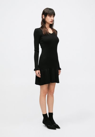 Čierne rebrované pletené šaty s dlhými rukávmi, priliehavým živôtikom a rozšíreným spodkom. Ozdobené riasenými manžetami, doplnené čiernymi špicatými topánkami.