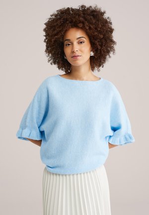 Kvinde med krøllet hår iført sølv hoops-øreringe, lyseblå kortærmet sweater og hvid plisseret nederdel, stående foran en neutral baggrund.