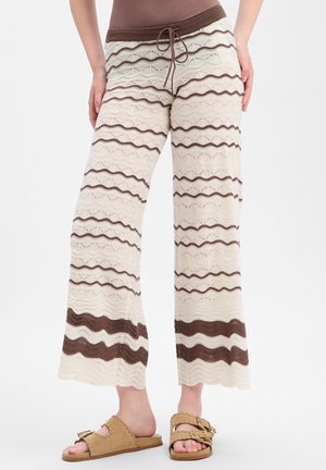 Pantalon en maille beige et marron à jambes larges avec des rayures horizontales ondulées, associé à des sandales à boucle ouvertes couleur cuir sur fond blanc.