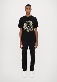 Black t-shirt featuring a circular graphic with white text and yellow floral accents, paired with black pants and white sneakers.
