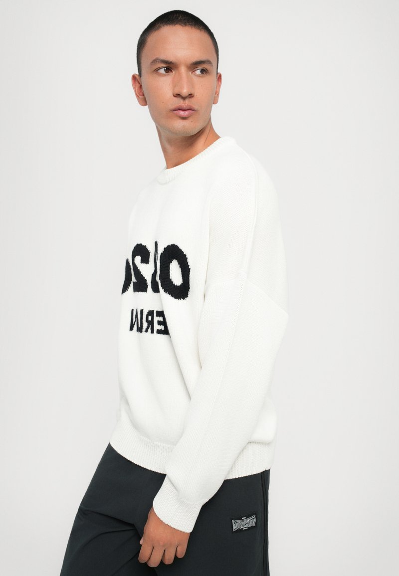 Pull en maille blanc avec un design de texte noir "OSLO" sur le devant, avec une coupe décontractée et des poignets côtelés. Porté avec un pantalon sombre.