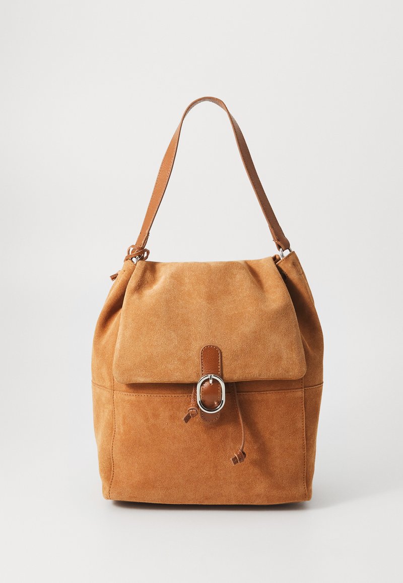 Tan suede shoulder bag with a fold-over flap, brown leather accents, and a silver hardware ring. Features a drawstring closure and soft texture.