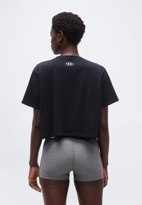 Άτομο που φοράει μαύρο crop T-shirt με λευκό λογότυπο και γκρι εφαρμοστά σορτς, όρθιο με την πλάτη να κοιτάζει προς την κάμερα σε απλό λευκό φόντο.