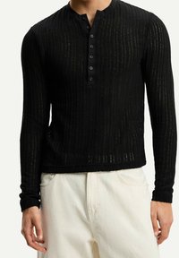 Camisa henley de manga larga negra con textura acanalada, que presenta un placket de botones y un cuello redondo. Combinada con pantalones de color claro.