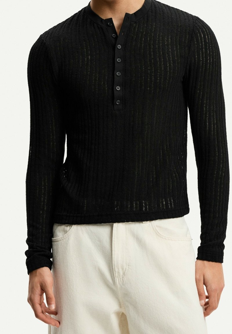 Camisa henley de manga larga negra con textura acanalada, que presenta un placket de botones y un cuello redondo. Combinada con pantalones de color claro.