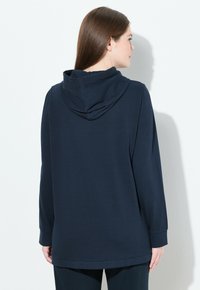 Donna con lunghi capelli castani che indossa un felpa blu navy a maniche lunghe, in piedi con la schiena rivolta verso la fotocamera contro un muro bianco.