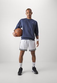 Atleta com uma sweatshirt azul-marinho e calções brancos com riscas pretas, segurando uma bola de basquetebol. Tecido texturizado, com calçado atlético e meias até ao tornozelo.