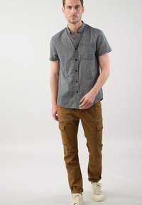 Korte mouwen, patroonhemd in donkere tinten; gecombineerd met kaki cargo shorts met ritszakken. Model staat met een neutrale uitdrukking.