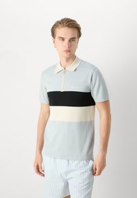 Polo em azul claro, com uma faixa horizontal preta e creme, fecho em zíper e logotipo bordado no peito. Mangas curtas.