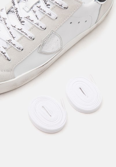 Baskets blanches et grises avec un texte manuscrit noir sur les lacets, à côté de deux lacets blancs enroulés sur un fond blanc.